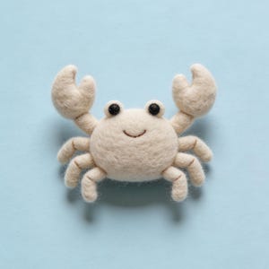 Puede incluir: Un cangrejo de fieltro blanco roto hecho a mano, con ojos negros y una boca sonriente. El cangrejo tiene dos grandes pinzas y ocho patas. Está colocado sobre un fondo azul claro, creando una estética caprichosa y juguetona.