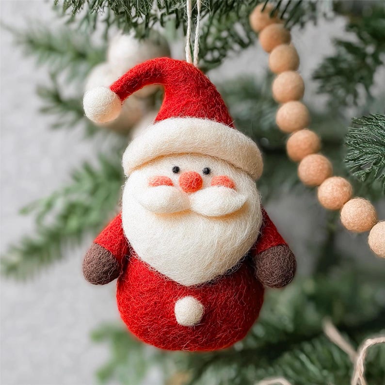 Pode incluir: Um enfeite de Natal artesanal em forma de Papai Noel. O enfeite apresenta um terno e chap&eacute;u de feltro vermelho, barba e acabamento brancos e um pequeno nariz laranja. Est&aacute; pendurado em uma &aacute;rvore de Natal com agulhas verdes e uma guirlanda de contas bege.