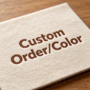May include: A close-up of a beige felt square with the words "Custom Order/Color" embossed in brown. The felt has a soft, textured appearance. The text is centered and clearly visible.
