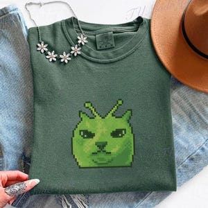 May include: A green crew neck sweatshirt featuring a pixelated green alien cat graphic. The sweatshirt is paired with blue jeans, a brown hat, and a floral necklace. The image is taken on a white surface.