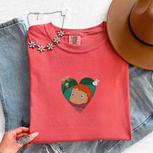 May include: Coral-colored t-shirt featuring a heart-shaped graphic with a cartoon character, strawberries, and a rabbit. The shirt is paired with blue jeans, a brown hat, and a floral necklace. The shirt has a round neck and short sleeves.
