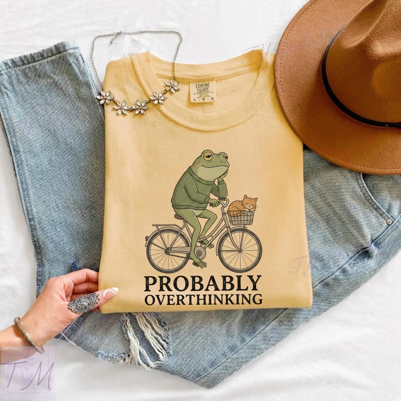 Op de afbeelding: Lichtgele t-shirt met een afbeelding van een kikker op een fiets met een kat in de mand. De tekst "PROBABLY OVERTHINKING" staat eronder. Het shirt ligt op een blauwe spijkerbroek.
