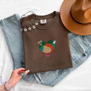 May include: A brown t-shirt featuring a heart-shaped graphic with a cartoon girl, strawberries, and a white rabbit. The shirt is folded on top of blue jeans, with a brown hat and a floral necklace.