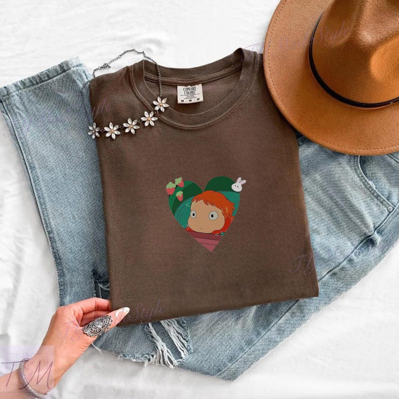 May include: A brown t-shirt featuring a heart-shaped graphic with a cartoon girl, strawberries, and a white rabbit. The shirt is folded on top of blue jeans, with a brown hat and a floral necklace.