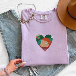 May include: A light purple t-shirt with a heart-shaped graphic featuring a cartoon character with red hair. The shirt is paired with blue jeans, a floral necklace, and a brown hat.