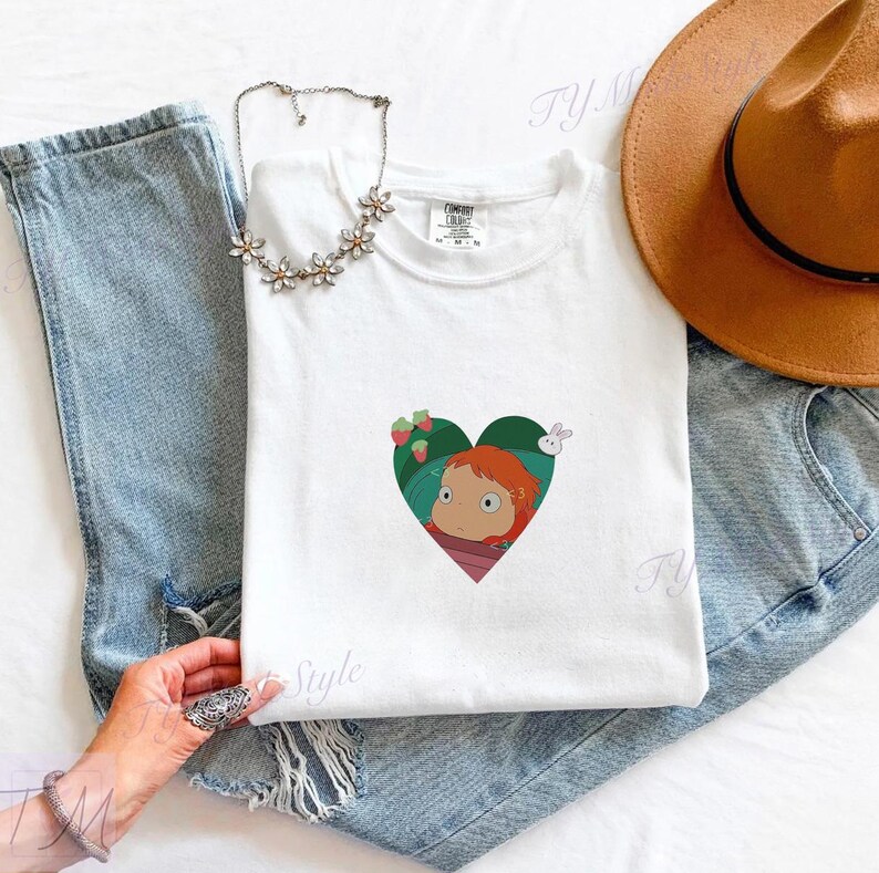 May include: White t-shirt featuring a heart-shaped graphic with a cartoon character, strawberries, and a rabbit. The shirt is paired with light blue jeans, a brown hat, and a floral necklace.