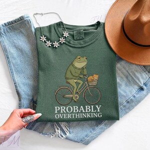 Op de afbeelding: Een groen t-shirt met een illustratie van een kikker op een fiets met een kat in de mand. De tekst "PROBABLY OVERTHINKING" staat eronder. Het t-shirt wordt gecombineerd met een blauwe spijkerbroek en een bruine hoed.