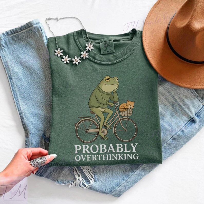 Op de afbeelding: Een groen t-shirt met een illustratie van een kikker op een fiets met een kat in de mand. De tekst "PROBABLY OVERTHINKING" staat eronder. Het t-shirt wordt gecombineerd met een blauwe spijkerbroek en een bruine hoed.