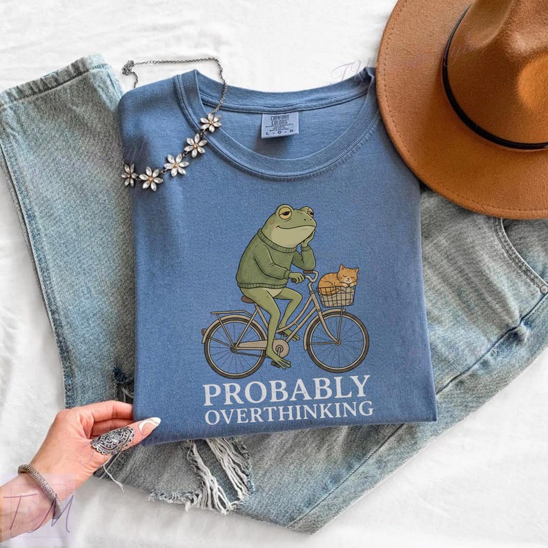 Op de afbeelding: Blauw T-shirt met een illustratie van een kikker op een fiets met een kat in de mand. De tekst "PROBABLY OVERTHINKING" staat eronder. Een bruine hoed en jeans zijn ook zichtbaar. De afbeelding is gestyled op een witte ondergrond.