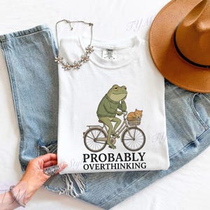 Op de afbeelding: Wit T-shirt met een cartoon kikker op een fiets en een kat in de mand. De kikker draagt een groene trui en denkt na. De tekst "PROBABLY OVERTHINKING" staat eronder. Het T-shirt ligt op een spijkerbroek.