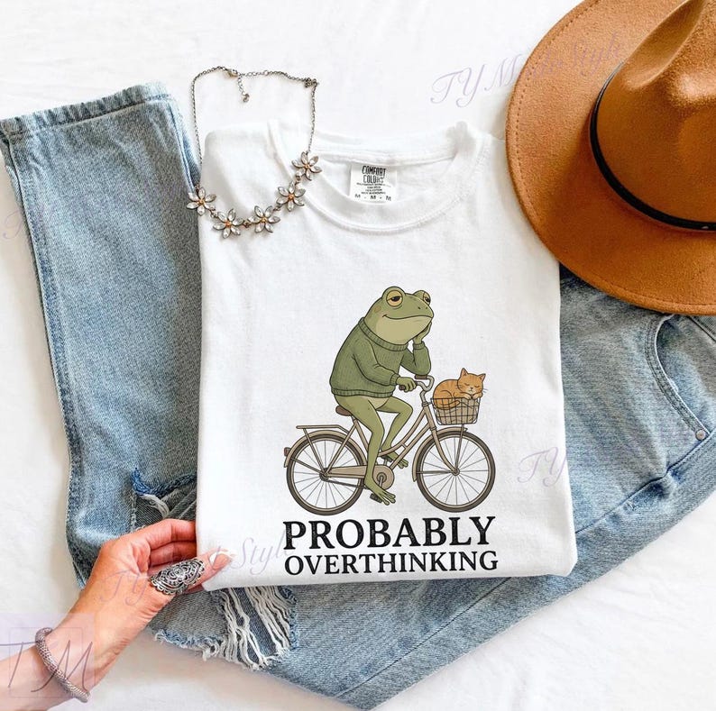 Op de afbeelding: Wit T-shirt met een cartoon kikker op een fiets en een kat in de mand. De kikker draagt een groene trui en denkt na. De tekst "PROBABLY OVERTHINKING" staat eronder. Het T-shirt ligt op een spijkerbroek.