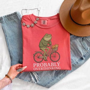Op de afbeelding: Koraalrood T-shirt met een kikker op een fiets en een kat in de mand. De kikker draagt een trui en lijkt na te denken. De tekst "PROBABLY OVERTHINKING" staat eronder. Het T-shirt wordt gecombineerd met een spijkerbroek en een bruine hoed.