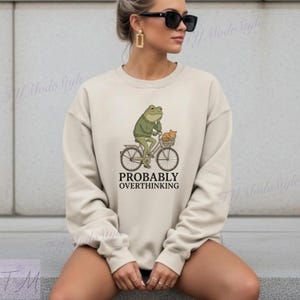 Op de afbeelding: Beige sweatshirt met een grappige afbeelding van een kikker op een fiets met een kat in een mand. De tekst "PROBABLY OVERTHINKING" staat eronder. De trui heeft een ronde hals en lange mouwen.