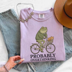 Op de afbeelding: Een lila T-shirt met een illustratie van een kikker op een fiets en een kat in de mand. De tekst "PROBABLY OVERTHINKING" staat eronder. Het T-shirt heeft een ronde hals en korte mouwen.
