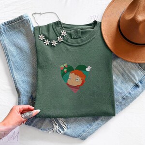 May include: A green crew neck t-shirt featuring a heart-shaped graphic with a cartoon character, strawberries, and a rabbit. The shirt is paired with blue jeans, a brown hat, and a floral necklace. The shirt is folded on a white surface.
