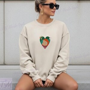 May include: Beige crewneck sweatshirt with a heart-shaped graphic. The green heart features a cartoon character with red hair and a pink scarf. The sweatshirt is a neutral color and has long sleeves.