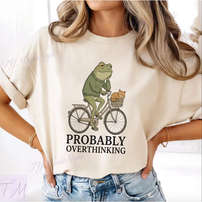 Op de afbeelding: Cr&egrave;mekleurig T-shirt met een illustratie van een kikker in een groene trui op een fiets met een kat in de mand. De tekst luidt "PROBABLY OVERTHINKING".