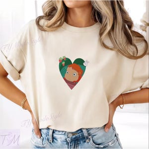 May include: Cream-colored t-shirt featuring a heart-shaped graphic with a cartoon character with red hair, strawberries, and a white rabbit. The shirt has short sleeves and a relaxed fit.