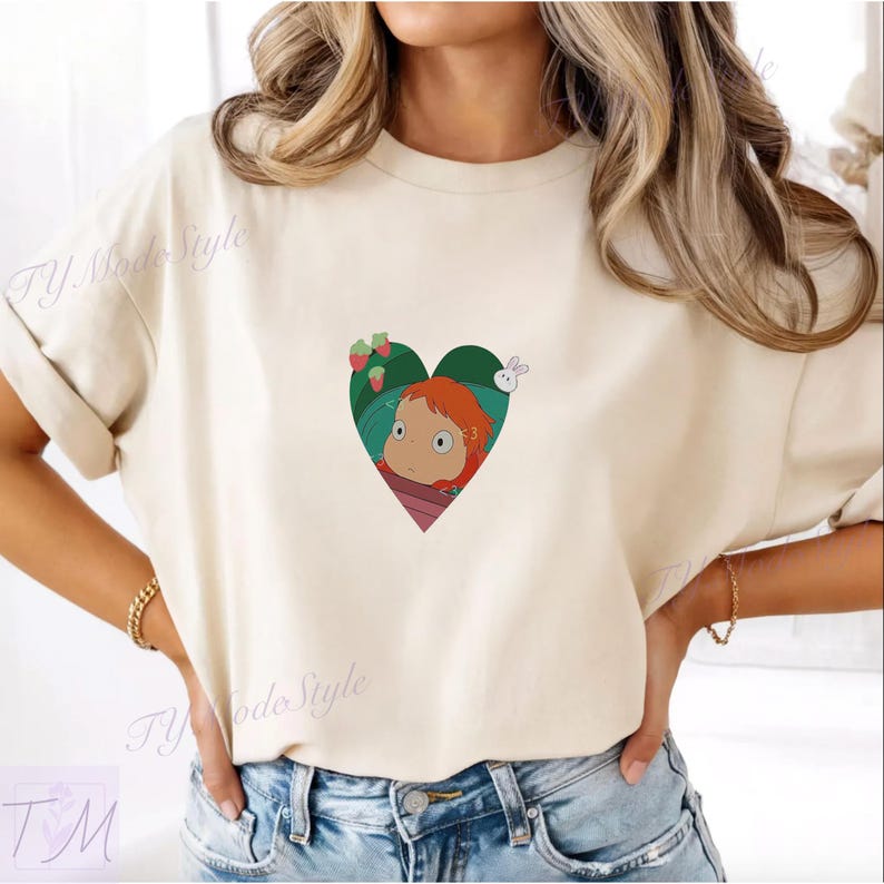 May include: Cream-colored t-shirt featuring a heart-shaped graphic with a cartoon character with red hair, strawberries, and a white rabbit. The shirt has short sleeves and a relaxed fit.