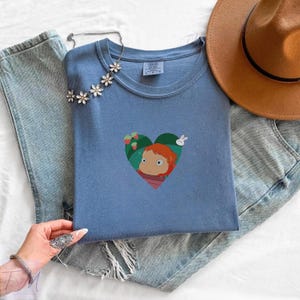 May include: A light blue t-shirt featuring a heart-shaped graphic with a cartoon character, strawberries, and a rabbit. The shirt is folded on top of a pair of light blue jeans. A floral necklace and a brown hat are also visible.