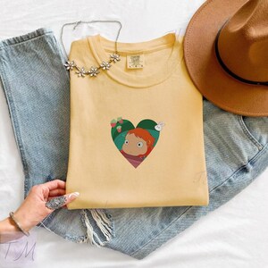 May include: A light yellow t-shirt featuring a heart-shaped graphic with a cartoon character, strawberries, and a rabbit. The shirt is laid on top of blue jeans, with a brown hat and a silver necklace.