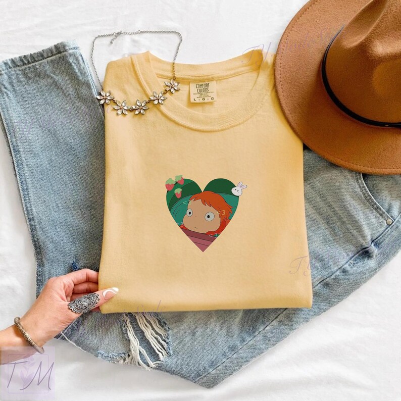 May include: A light yellow t-shirt featuring a heart-shaped graphic with a cartoon character, strawberries, and a rabbit. The shirt is laid on top of blue jeans, with a brown hat and a silver necklace.