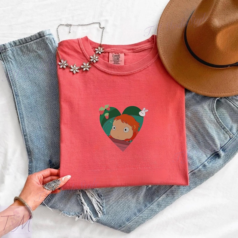 May include: Coral-colored t-shirt featuring a heart-shaped graphic with a cartoon character, strawberries, and a rabbit. The shirt is paired with blue jeans, a brown hat, and a floral necklace. The shirt has a round neck and short sleeves.