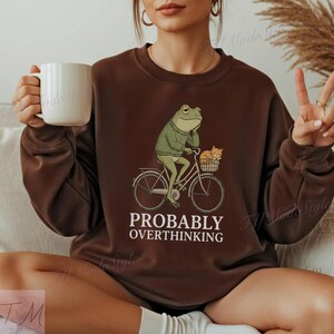 Op de afbeelding: Bruin sweatshirt met een illustratie van een kikker op een fiets met een kat in de mand. De tekst "PROBABLY OVERTHINKING" staat eronder gedrukt. Het sweatshirt is effen bruin van kleur.