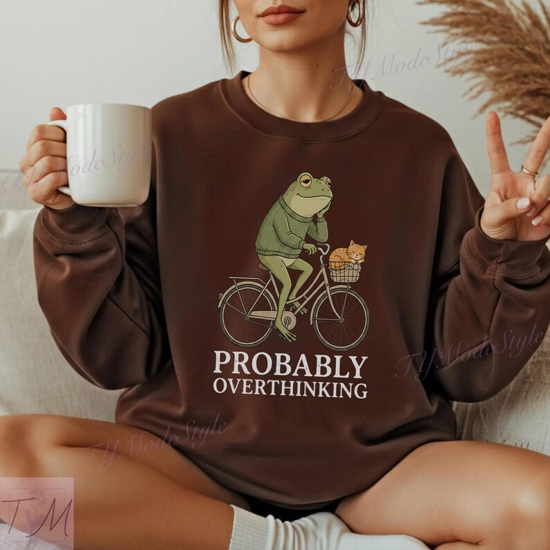 Op de afbeelding: Bruin sweatshirt met een illustratie van een kikker op een fiets met een kat in de mand. De tekst "PROBABLY OVERTHINKING" staat eronder gedrukt. Het sweatshirt is effen bruin van kleur.