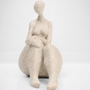 May include: A beige, textured sculpture of a seated figure with arms crossed over the knees. The figure has a smooth, rounded head and a simple, minimalist design. The sculpture is on a white background.