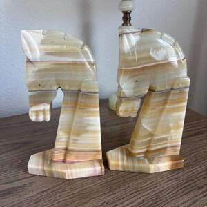 May include: A pair of horse head bookends carved from banded onyx. The bookends feature a cream, tan, and brown color palette. The horse heads are in a stylized, geometric design, with a rectangular base.