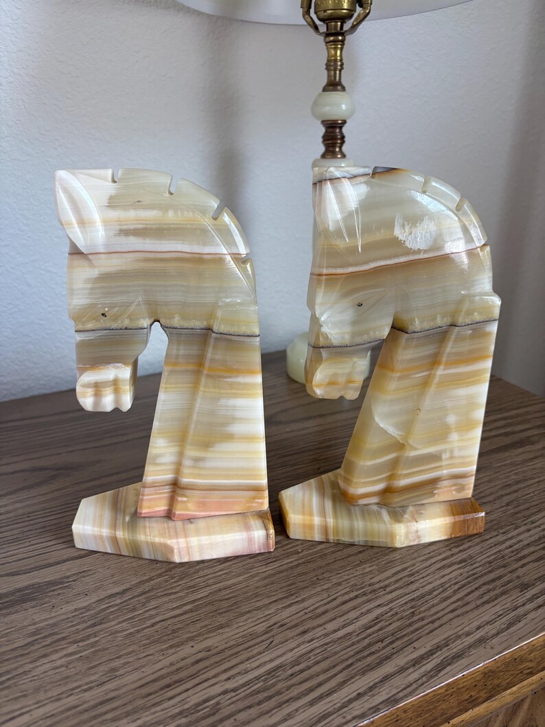May include: A pair of horse head bookends carved from banded onyx. The bookends feature a cream, tan, and brown color palette. The horse heads are in a stylized, geometric design, with a rectangular base.