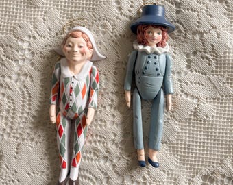 May include: Two decorative figurines. One is a Pierrot in a diamond pattern suit of red, white, and green. The other is a figure in a blue outfit with a matching hat and white ruffled collar. Both have painted faces and gold hanging loops.