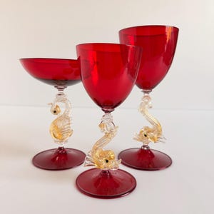May include: Three ruby red glass goblets with clear and gold-flecked stems shaped like seahorses. Each glass features a deep red bowl and a circular red base, with the central seahorse stem connecting the bowl to the base in a decorative design.