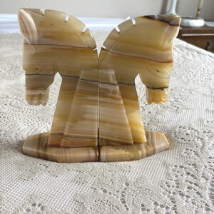 May include: A pair of onyx horse bookends. The bookends are carved from a light brown and tan stone, with a layered pattern. The horses are facing each other, with detailed manes and facial features. The bookends sit on a rectangular base.
