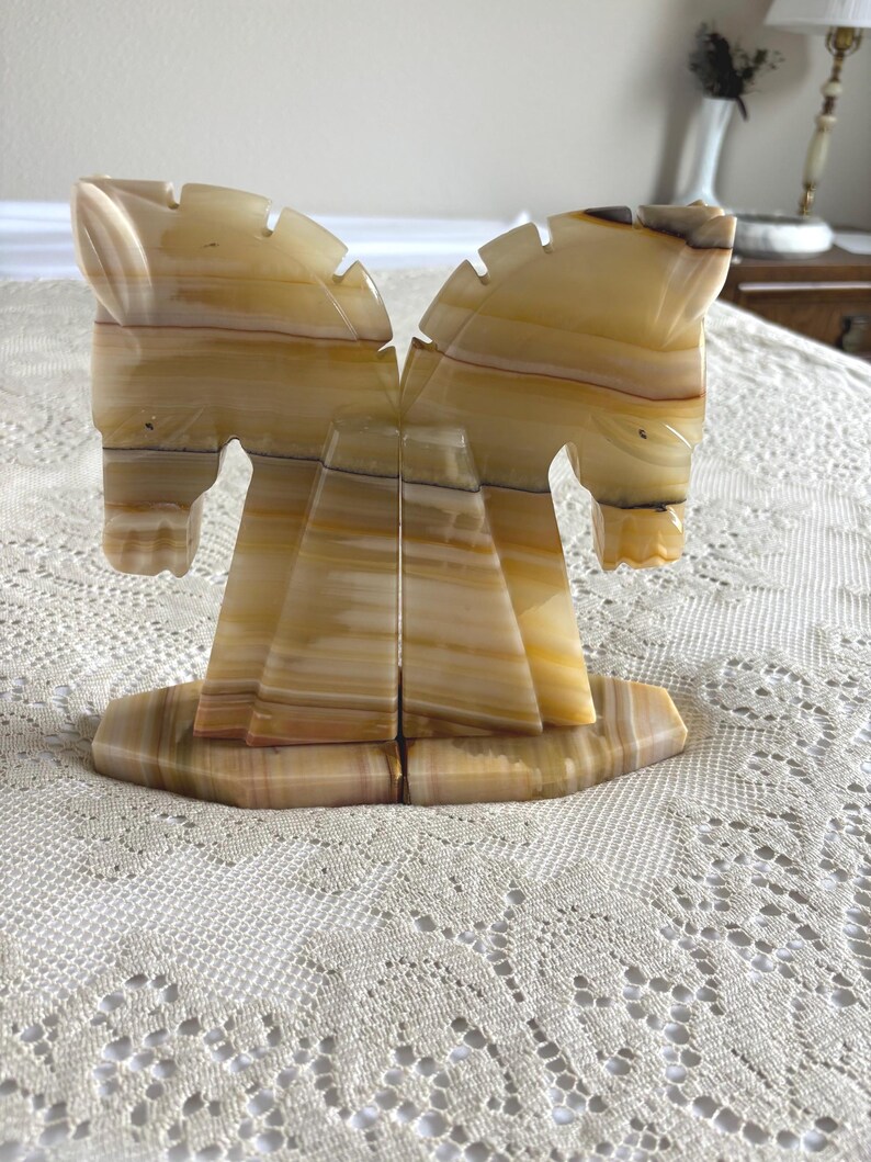 May include: A pair of onyx horse bookends. The bookends are carved from a light brown and tan stone, with a layered pattern. The horses are facing each other, with detailed manes and facial features. The bookends sit on a rectangular base.