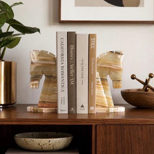 May include: A pair of horse-shaped bookends made of a light-colored, patterned stone. The bookends are holding several books with titles including "California Romantica" and "Still". The bookends are on a wooden surface.