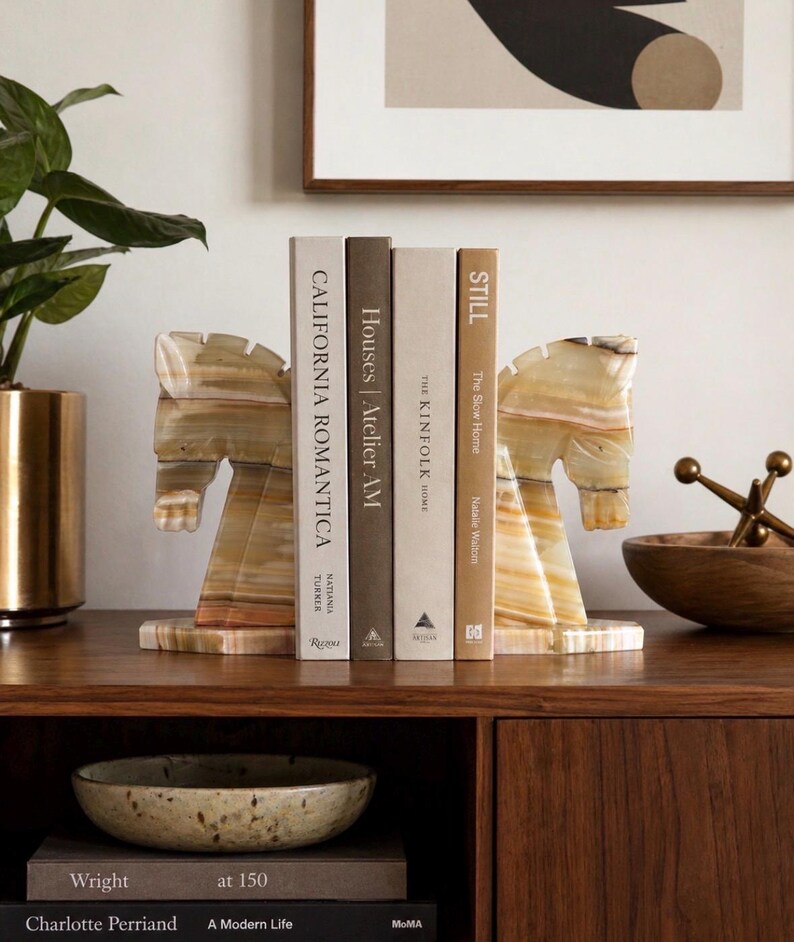 May include: A pair of horse-shaped bookends made of a light-colored, patterned stone. The bookends are holding several books with titles including "California Romantica" and "Still". The bookends are on a wooden surface.