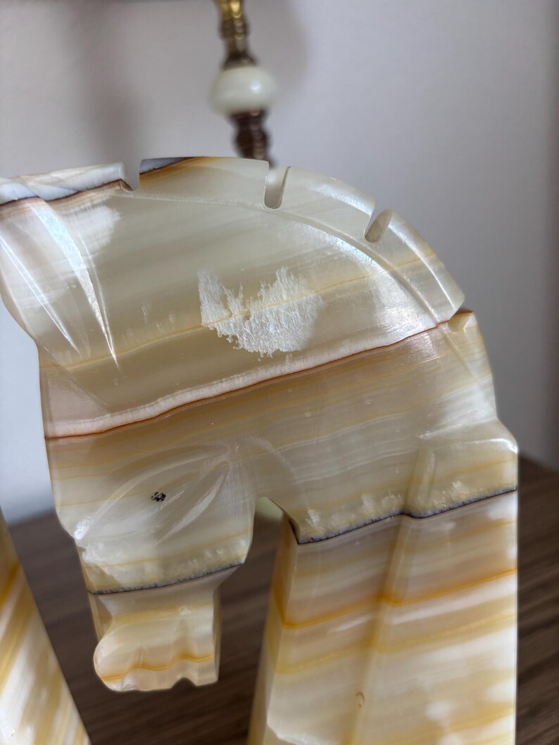 May include: Close-up of a carved onyx horse head sculpture. The sculpture features a cream, brown, and yellow color palette with visible veining. The horse head has a detailed mane and is part of a decorative art piece.