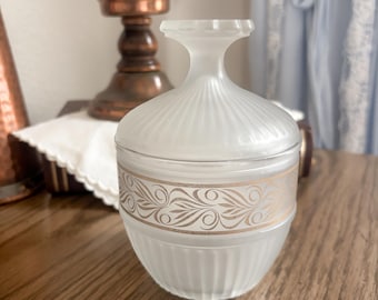 May include: A frosted glass jar with a lid, featuring a gold floral design around the middle. The jar has vertical ribbed detailing and a rounded base. A copper candlestick and a white candle are in the background.
