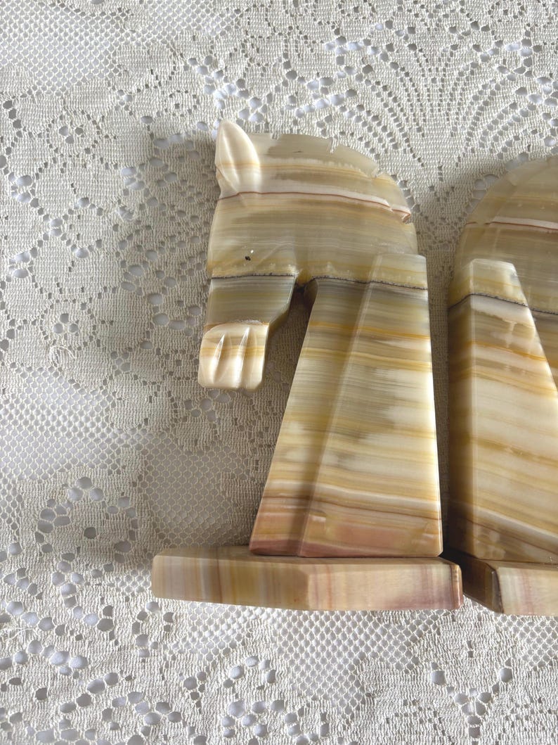 May include: A pair of decorative bookends carved from banded onyx. The bookends are shaped like horses, with tan, cream, and brown color variations. The horse heads are detailed with carved features.