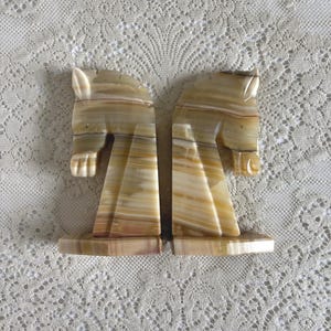 May include: Two onyx bookends, each carved in the shape of a horse's head. The bookends have a natural color palette of beige, brown, and cream, with a polished finish. The horse heads are facing opposite directions.