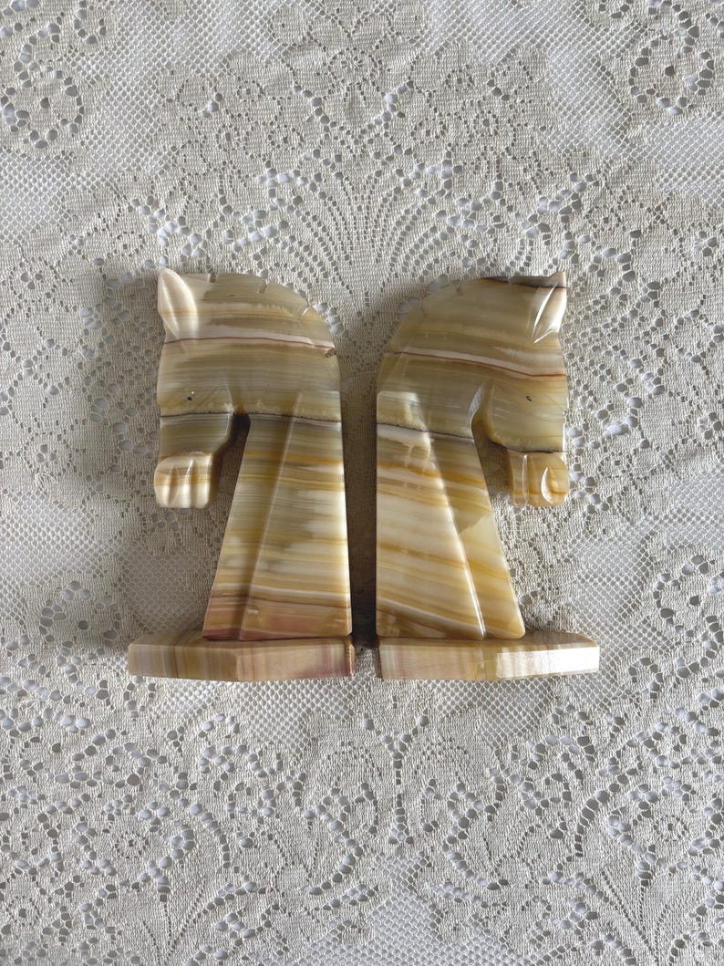 May include: Two onyx bookends, each carved in the shape of a horse's head. The bookends have a natural color palette of beige, brown, and cream, with a polished finish. The horse heads are facing opposite directions.