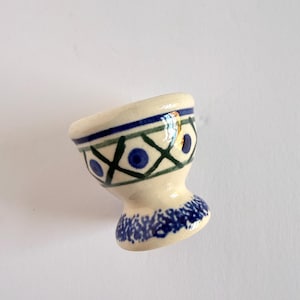 May include: A ceramic egg cup with a cream-colored base featuring a hand-painted pattern of dark green crisscross lines and blue dots. The rim is decorated with a blue band, and the pedestal base has a textured blue sponge-painted finish.