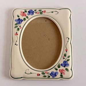 May include: A rectangular ceramic picture frame with an oval center opening. The frame is off-white with a dark green border and features hand-painted floral motifs in blue, pink, and green vines along the left and bottom edges.