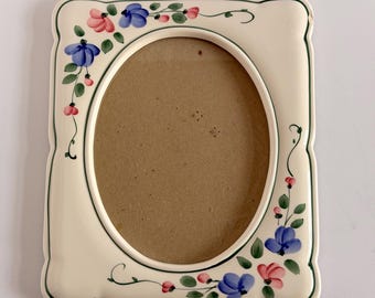 May include: A rectangular ceramic picture frame with an oval center opening. The frame is off-white with a dark green border and features hand-painted floral motifs in blue, pink, and green vines along the left and bottom edges.