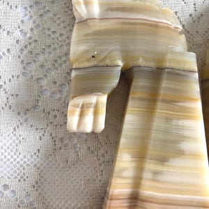 May include: A carved onyx sculpture with a layered, striped pattern in shades of cream, tan, and light brown. The sculpture appears to be a stylized animal figure, possibly a cat or a similar creature, with a smooth, polished finish.