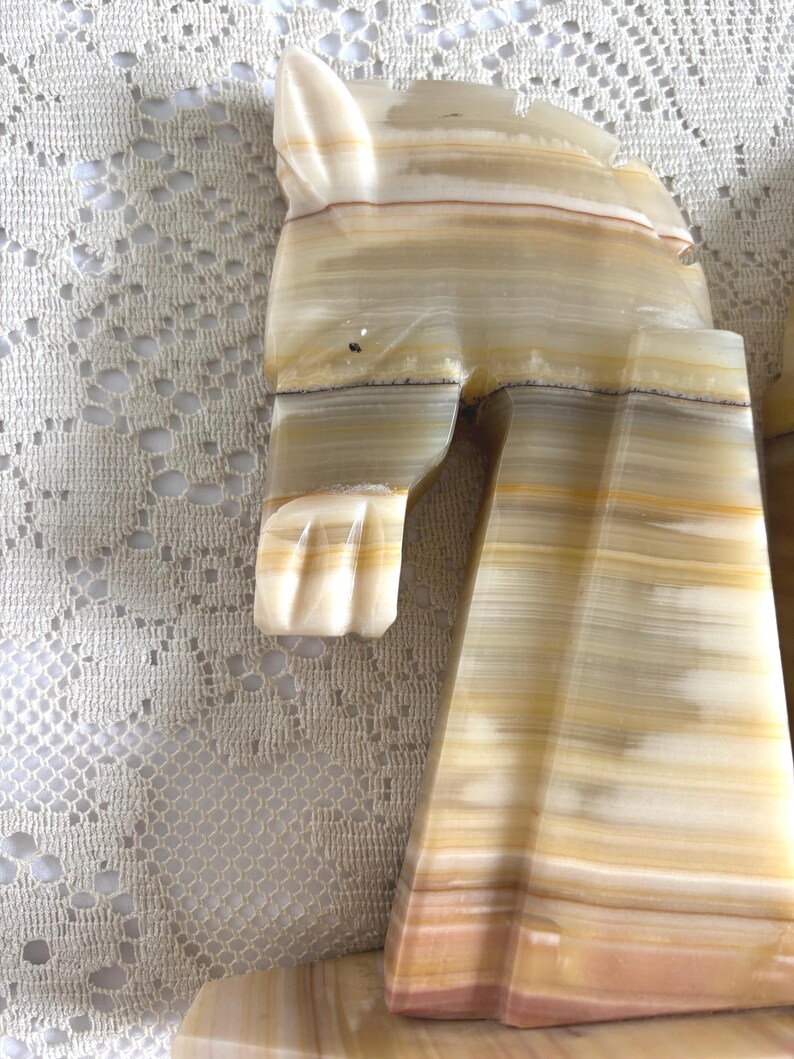 May include: A carved onyx sculpture with a layered, striped pattern in shades of cream, tan, and light brown. The sculpture appears to be a stylized animal figure, possibly a cat or a similar creature, with a smooth, polished finish.
