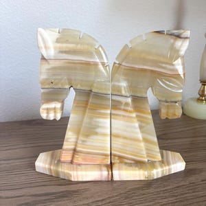 May include: A pair of onyx horse head bookends. The bookends are carved from a stone with beige, tan, and brown striations. The horse heads are facing each other, and the bases are a geometric shape.