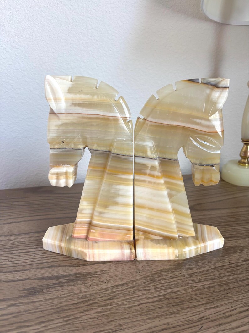 May include: A pair of onyx horse head bookends. The bookends are carved from a stone with beige, tan, and brown striations. The horse heads are facing each other, and the bases are a geometric shape.